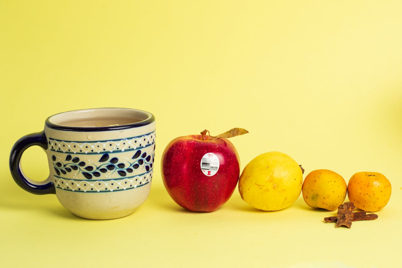 Colorful still life of ceramic mug, apple, and assorted fruits on yellow background.